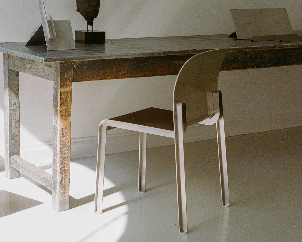 The Rivet Chair in brushed aluminium with a warm timber seat, drawn up to a weathered zinc-topped work table in a sunlit white room — Courtesy FRAMA